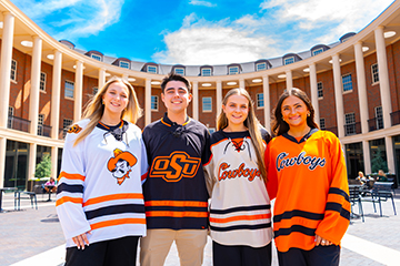 Four Oklahoma State University students wearing OSU hockey jerseys stand side by side outdoors in front of the Spears School of Business colonnade, posed for a promotional campus portrait.