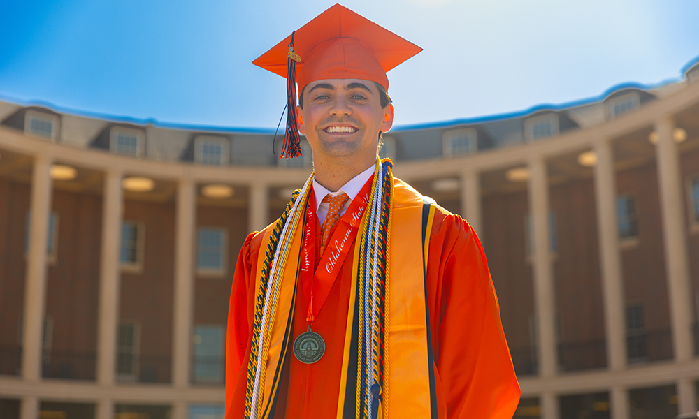 A graduate wearing an orange cap and gown with honor cords and a medallion stands outdoors in front of a curved colonnade building on the Oklahoma State University campus for a formal graduation portrait.