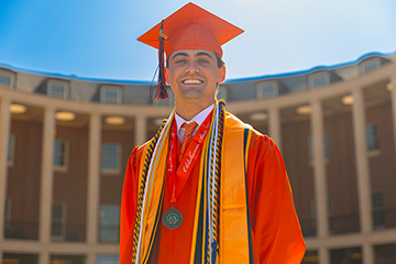 A graduate wearing an orange cap and gown with honor cords and a medallion stands outdoors in front of a curved colonnade building on the Oklahoma State University campus for a formal graduation portrait.