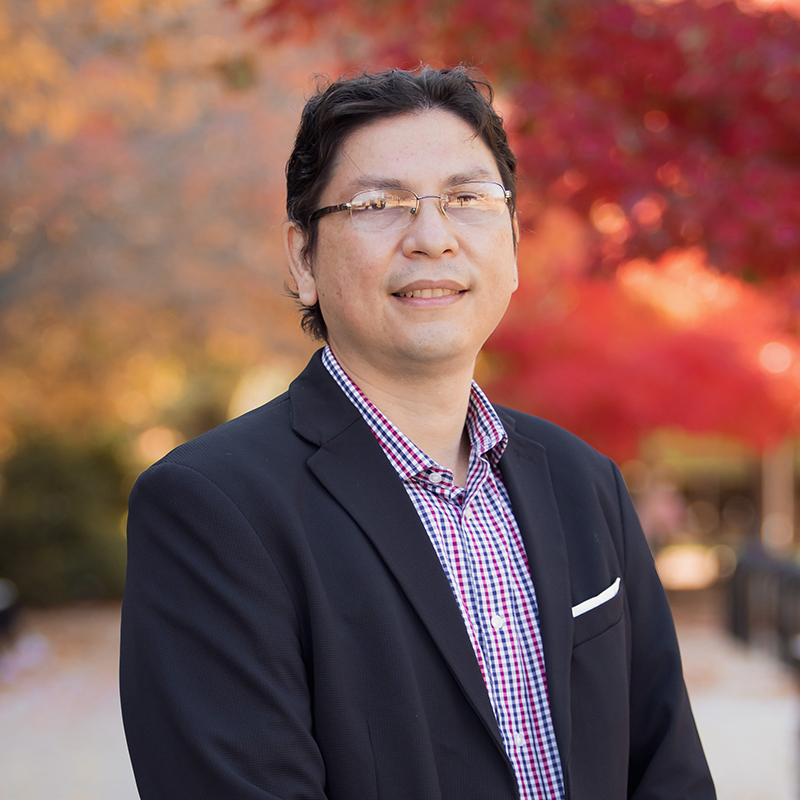 Jorge Arteaga-Fonseca wears eyeglasses and business attire as he smiles at the camera. Autumn leaves provide a scenic backdrop.