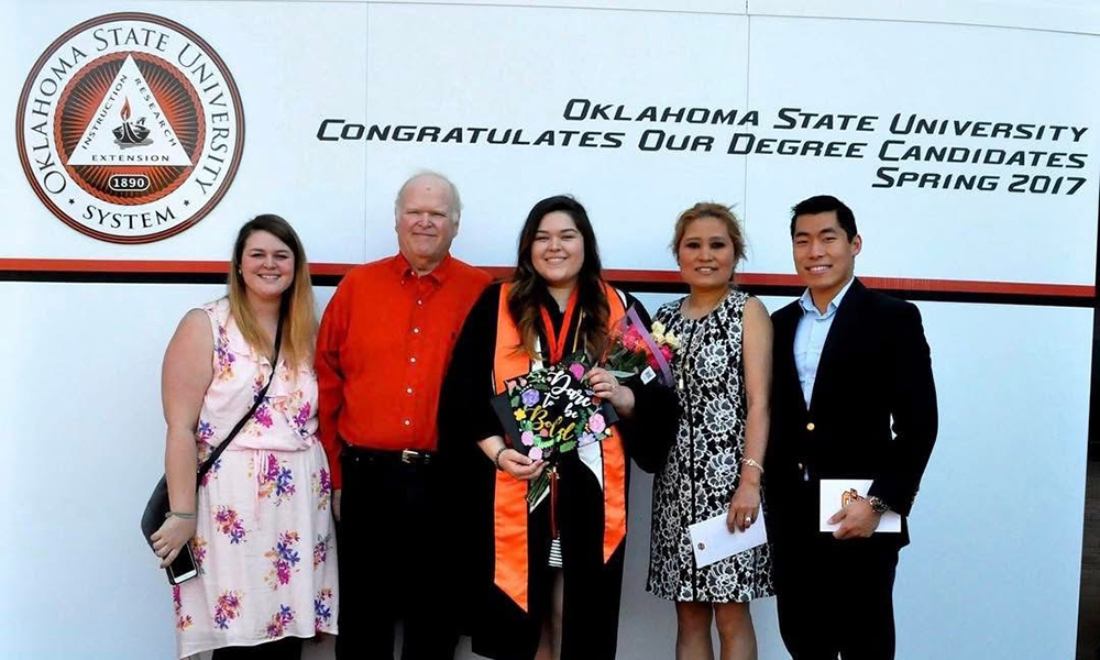 A woman celebrates college graduation with her family. She wears a black gown and holds a cap decorated with the phrase, "Dare to be bold."