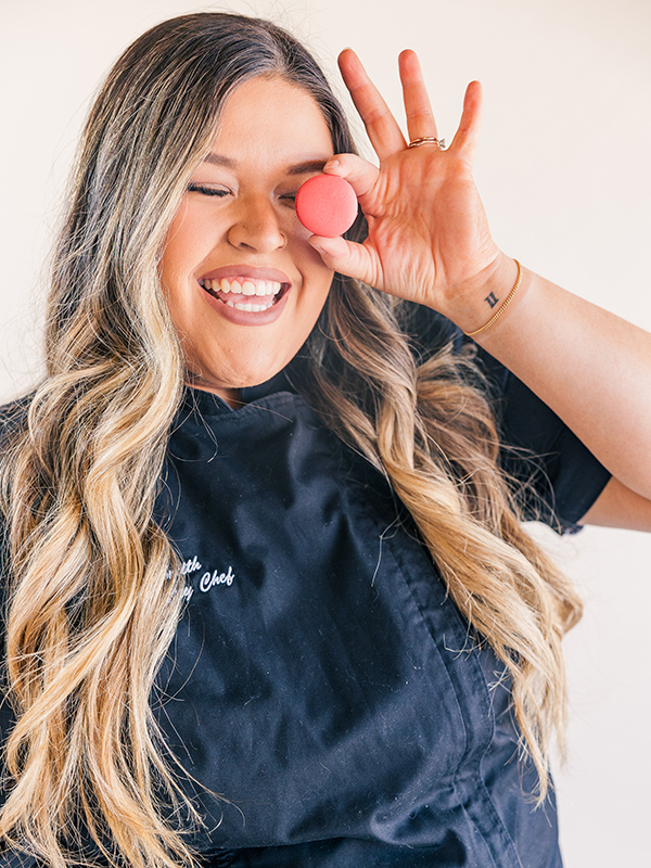 A woman smiles while holding up a red macaron. The woman has long, brown hair with blonde highlights. Her black shirt displays her title of "chef" in white, cursive letters.
