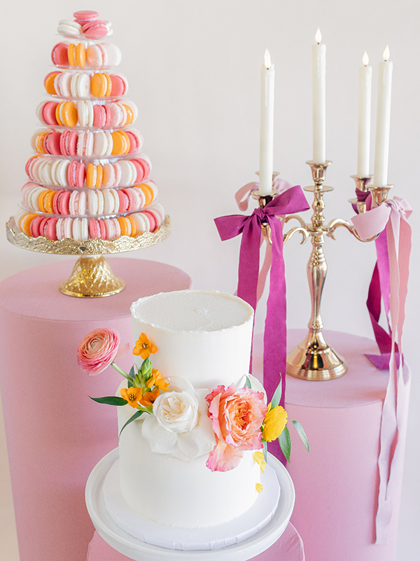 A tower of pastel macarons, a white wedding cake and a gold candelabra sit on pink platforms of various heights.