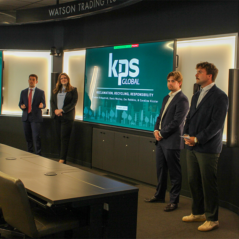 A team of four MBA students in business attire speaking via video chat with a group of executives from KPS Global in a modern classroom setting.