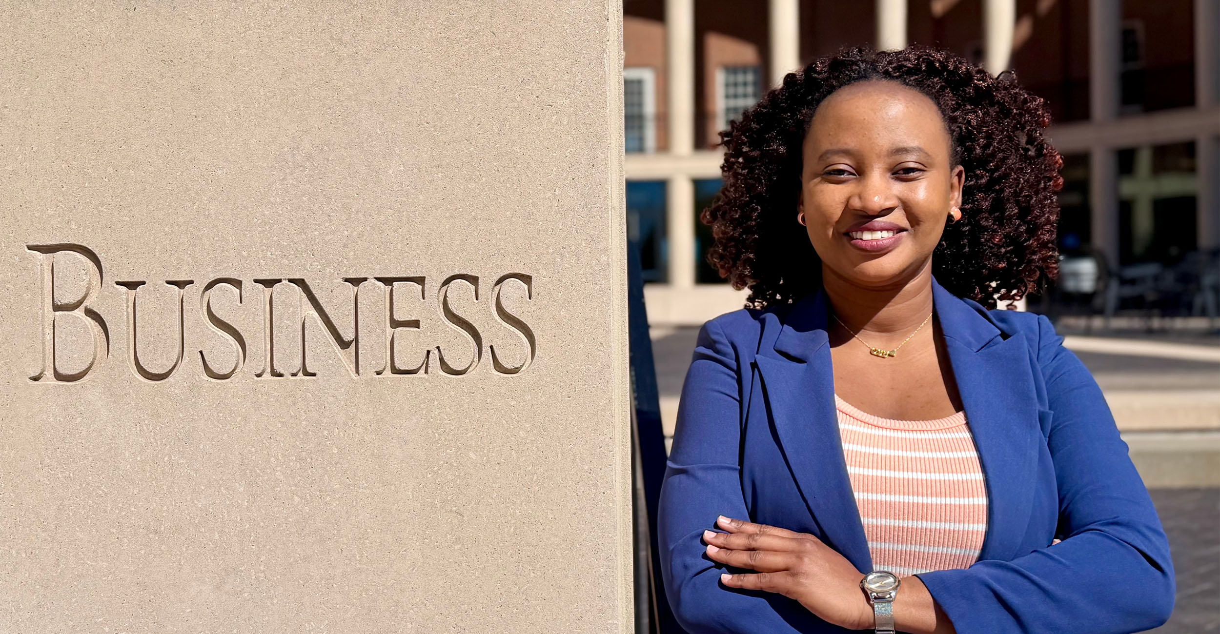 Person standing with arms crossed beside a stone pillar engraved with the word “Business” outside a university building, with campus architecture visible in the background.