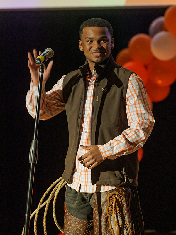 A young man wears a Western-inspired outfit as he speaks into a microphone on stage.
