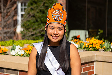 A young Chickasaw woman wears a beaded orange crown, a blue dress and a pageant sash. She waves and smiles.