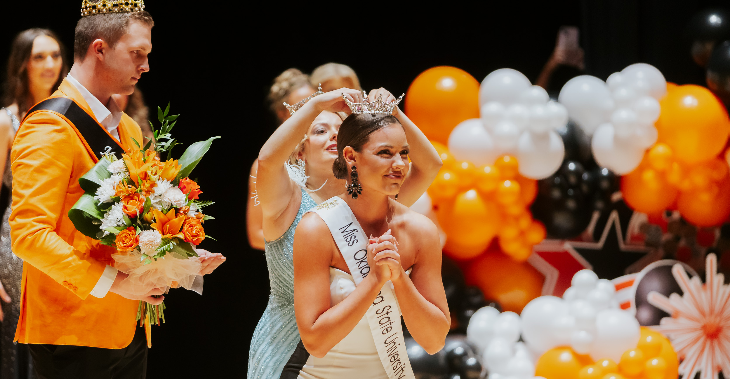 A young woman with brown hair wears a white gown and a sash as she stands on a stage. A young, blonde woman places a sparkly crown on the head of the woman in the white gown. Orange, black and white balloons decorate the stage, creating scenery fit for a pageant.