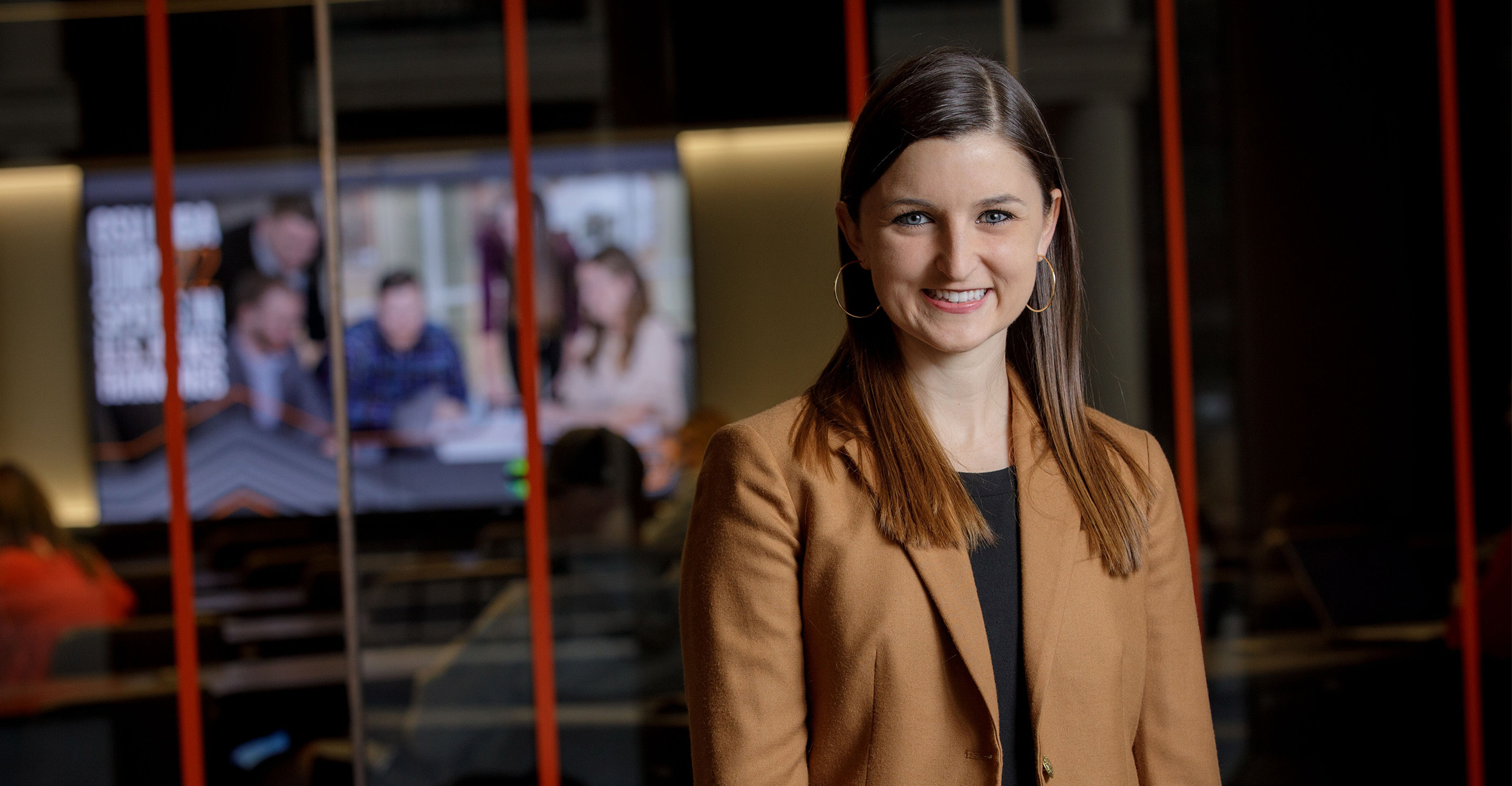 A person wearing a tan blazer stands in a modern indoor workspace with glass walls and illuminated screens showing a group discussion in the background, suggesting a professional media, newsroom, or presentation environment.