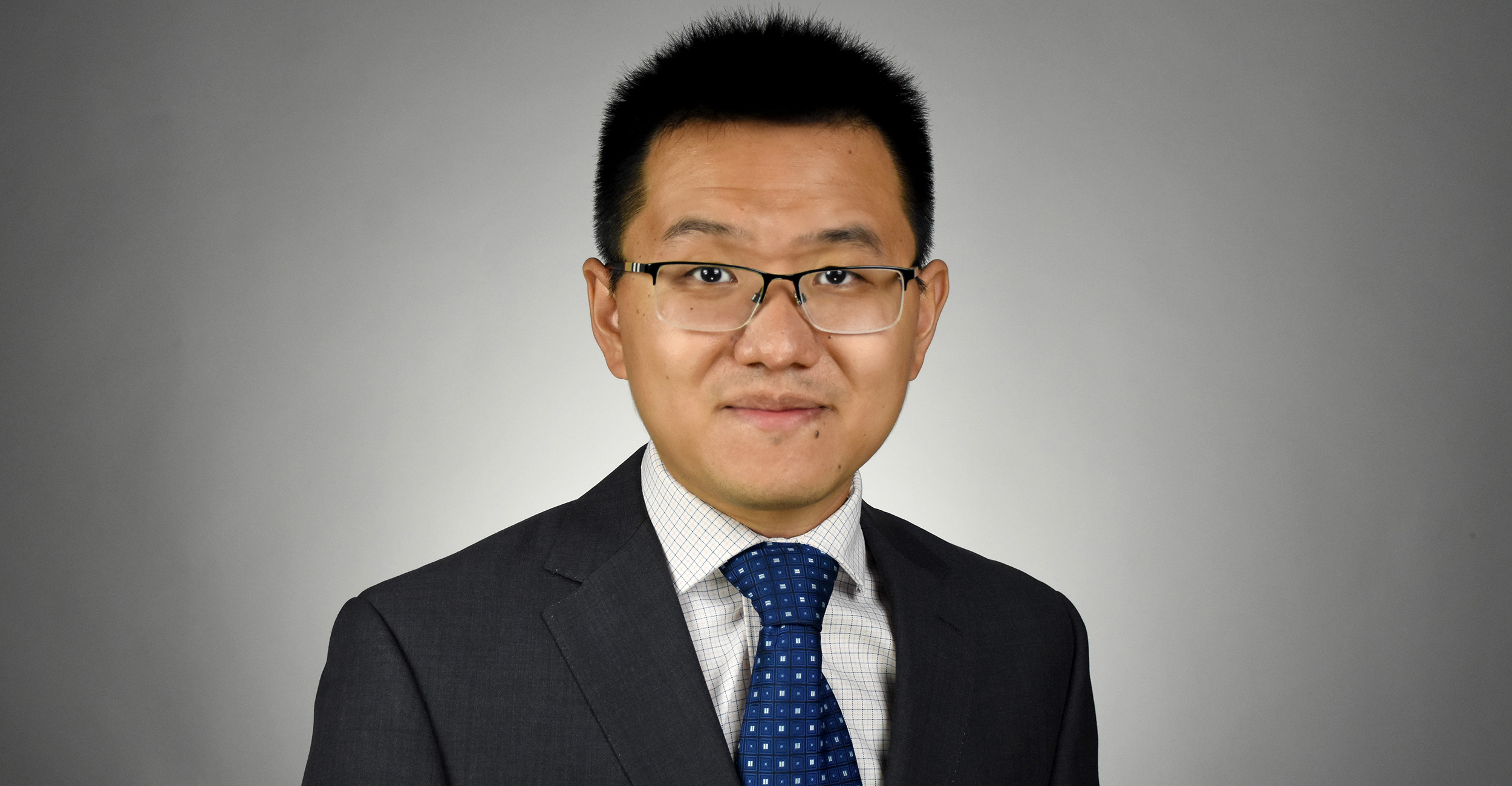 Person wearing a dark gray suit, white dress shirt, and blue patterned tie, posed against a plain gray background.