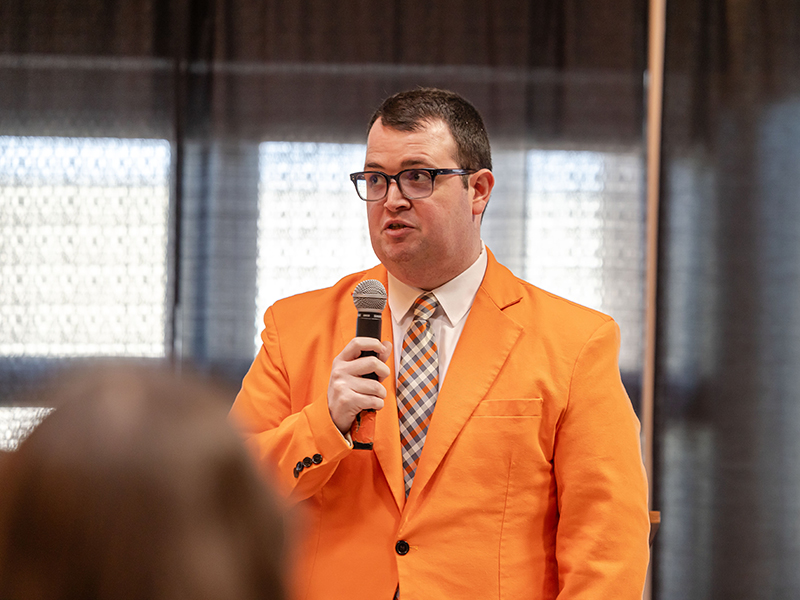 Sam Morse, wearing an orange jacket, speaks into a microphone.