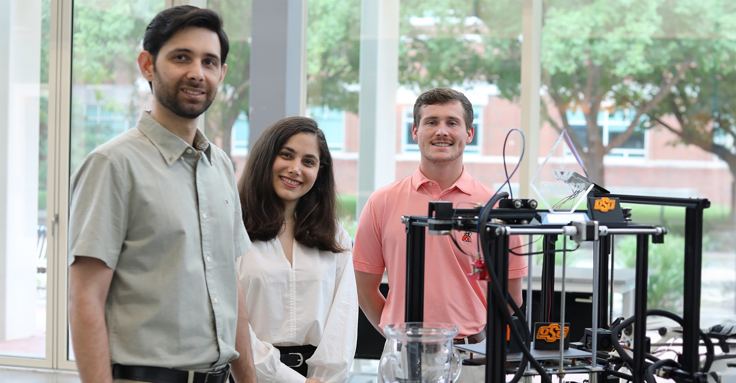 Three students stand beside a 3D printer and lab equipment inside a campus workspace, representing collaborative student research and hands‑on engineering work.