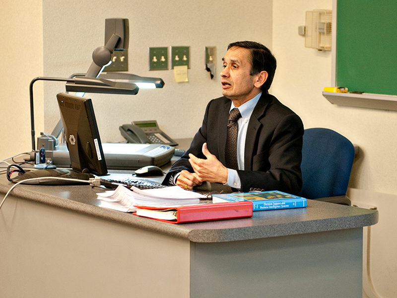 A man sits at a desk and lectures to his class.