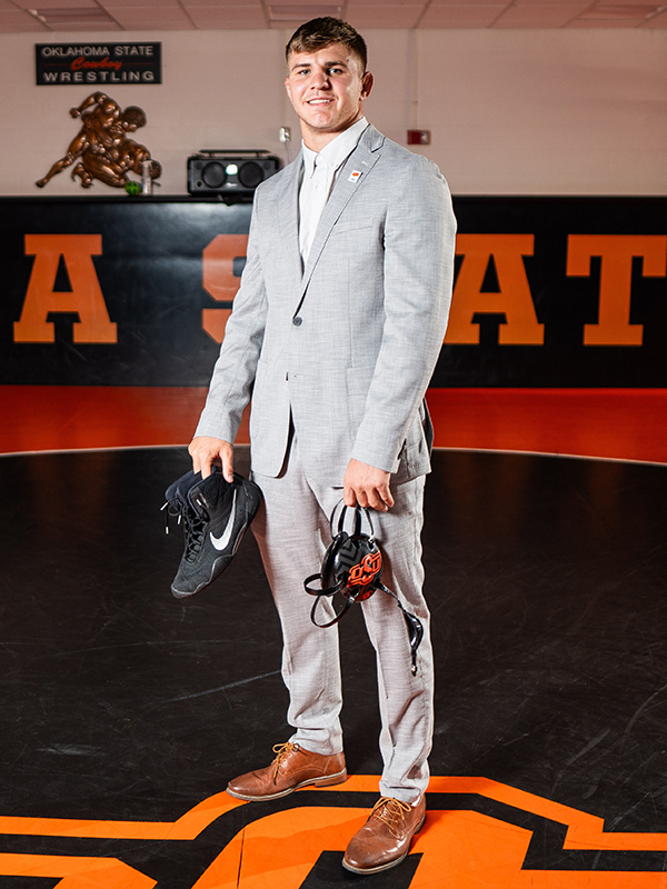 Wearing a business suit and MBA pin, Luke Surber holds his headgear and wrestling shoes in OSU's wrestling room.