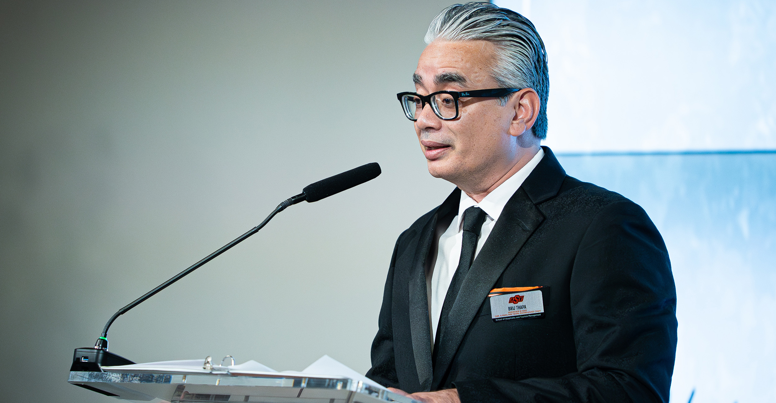 A man with gray hair, wearing a black suit and tie, stands at a clear podium with papers on it. He is speaking into a microphone attached to the podium. The background features a light-colored wall and a large window or screen.