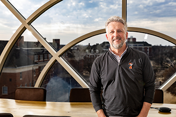 Person wearing a dark quarter‑zip pullover stands indoors in front of a large arched window, with a university campus visible outside, conveying a professional portrait in an academic setting.