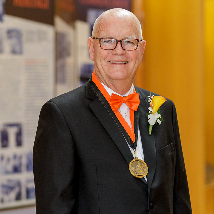 Portrait of a person wearing glasses, a black tuxedo with an orange bow tie, and a medal, posing indoors in front of display panels, highlighting a formal recognition or award event.