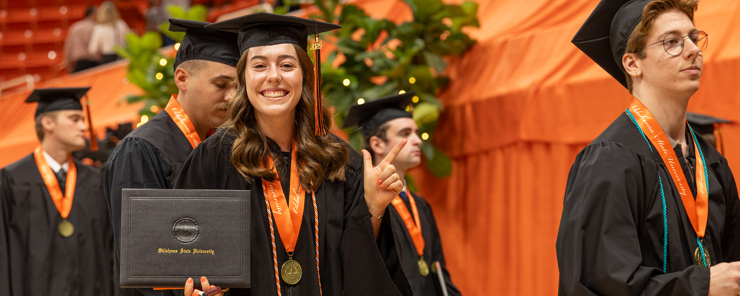 Graduates in black caps and gowns walk in procession during a commencement ceremony inside an arena decorated in orange. One graduate in the foreground smiles while holding a diploma cover, wearing an orange stole and medallion. Other graduates and greenery are visible in the background.