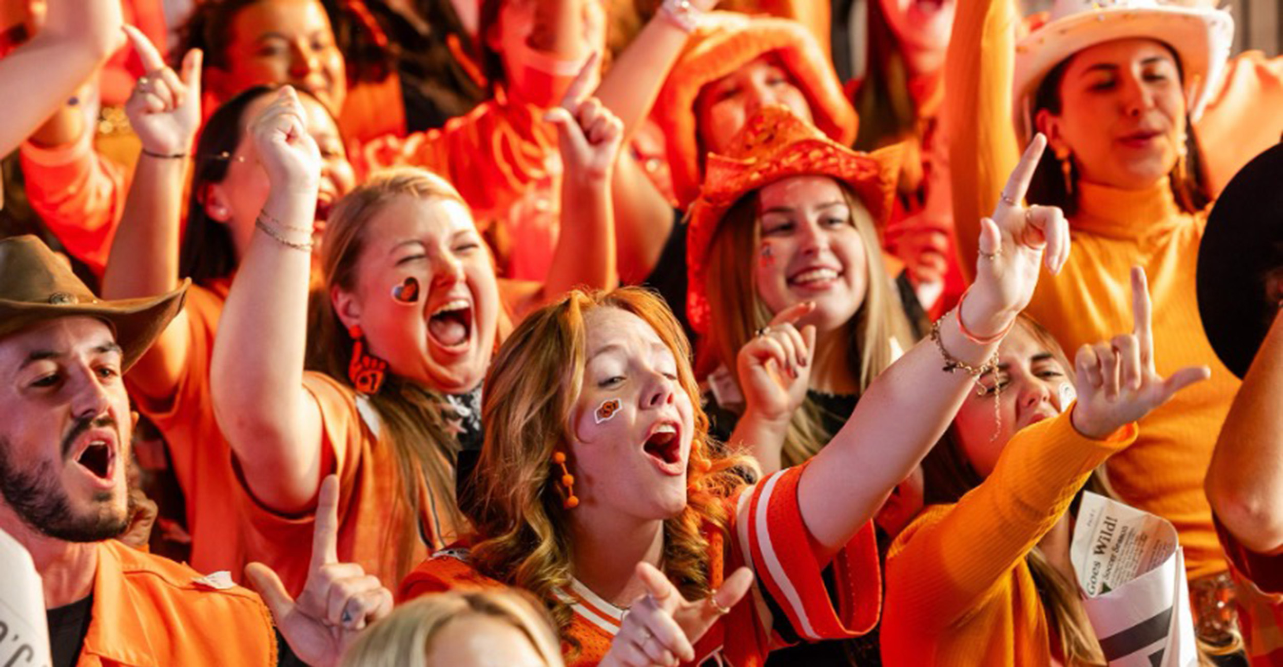 A crowd of people dressed in orange enthusiastically cheer with raised arms at a lively event, conveying excitement and school or team spirit.