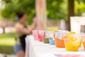 Clear plastic cups filled with brightly colored paint are lined along a table outdoors, with a person blurred in the background painting on a vertical surface, suggesting an outdoor art activity.