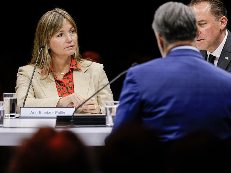Panelists sit at a conference table with microphones and name placards during a formal discussion or hearing, viewed from the audience perspective in a dimly lit venue.