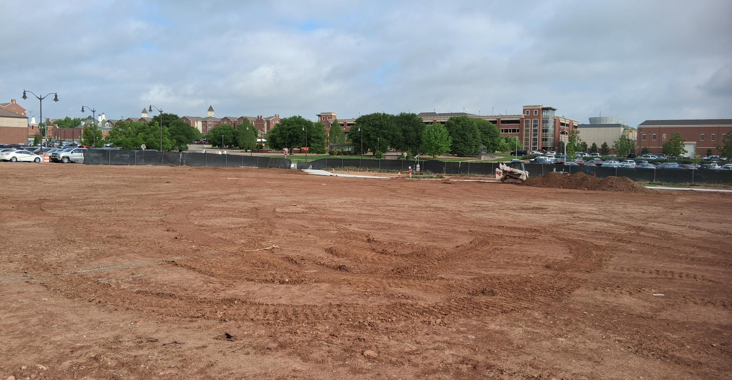 Color photograph of a cleared dirt construction site with fencing and equipment in the foreground and Oklahoma State University campus buildings and parking areas in the background under a cloudy sky.