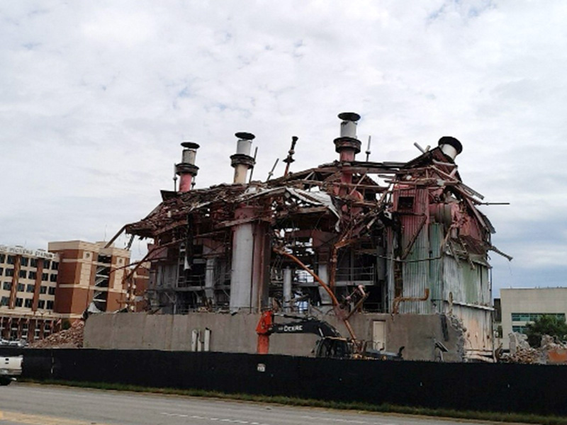 Color photograph of a partially demolished industrial power plant with exposed steel framework and smokestacks, surrounded by fencing and nearby campus buildings.