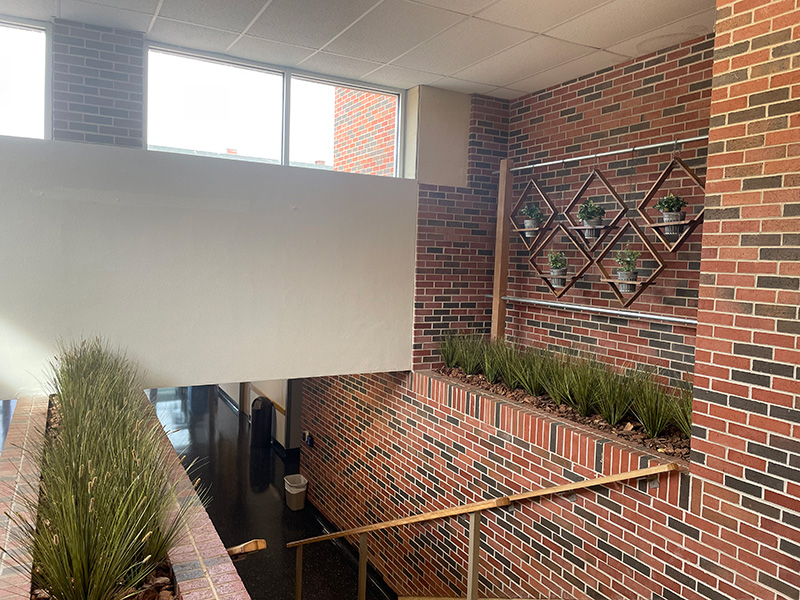 Color photograph of an indoor lobby featuring a brick half-wall with planted greenery and a series of colorful abstract paintings mounted high along the wall above a stairwell.