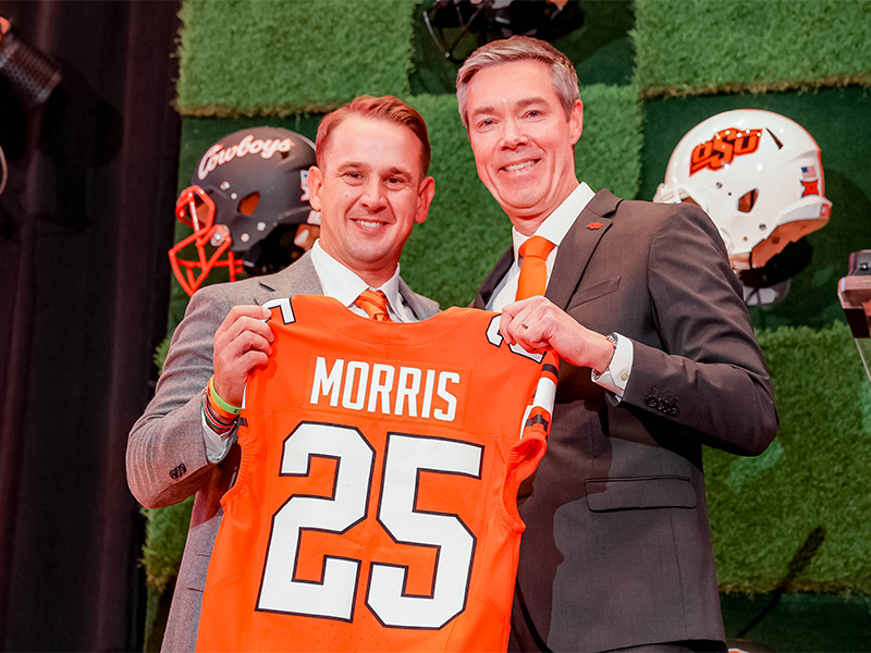 Two people in business attire stand on a stage holding up an orange football jersey with the name “Morris” and the number 25, with team helmets and sports‑themed decor in the background, indicating a formal athletic introduction or announcement.