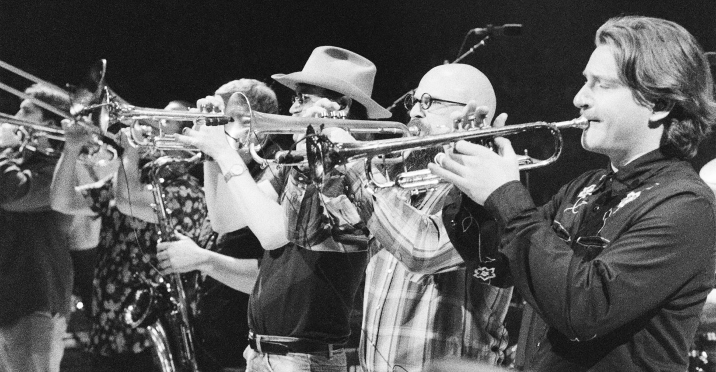 A black-and-white photograph shows a line of musicians standing on stage and playing brass instruments, including trumpets and trombones, during a live performance.