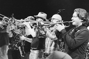 A black-and-white photograph shows a line of musicians standing on stage and playing brass instruments, including trumpets and trombones, during a live performance.