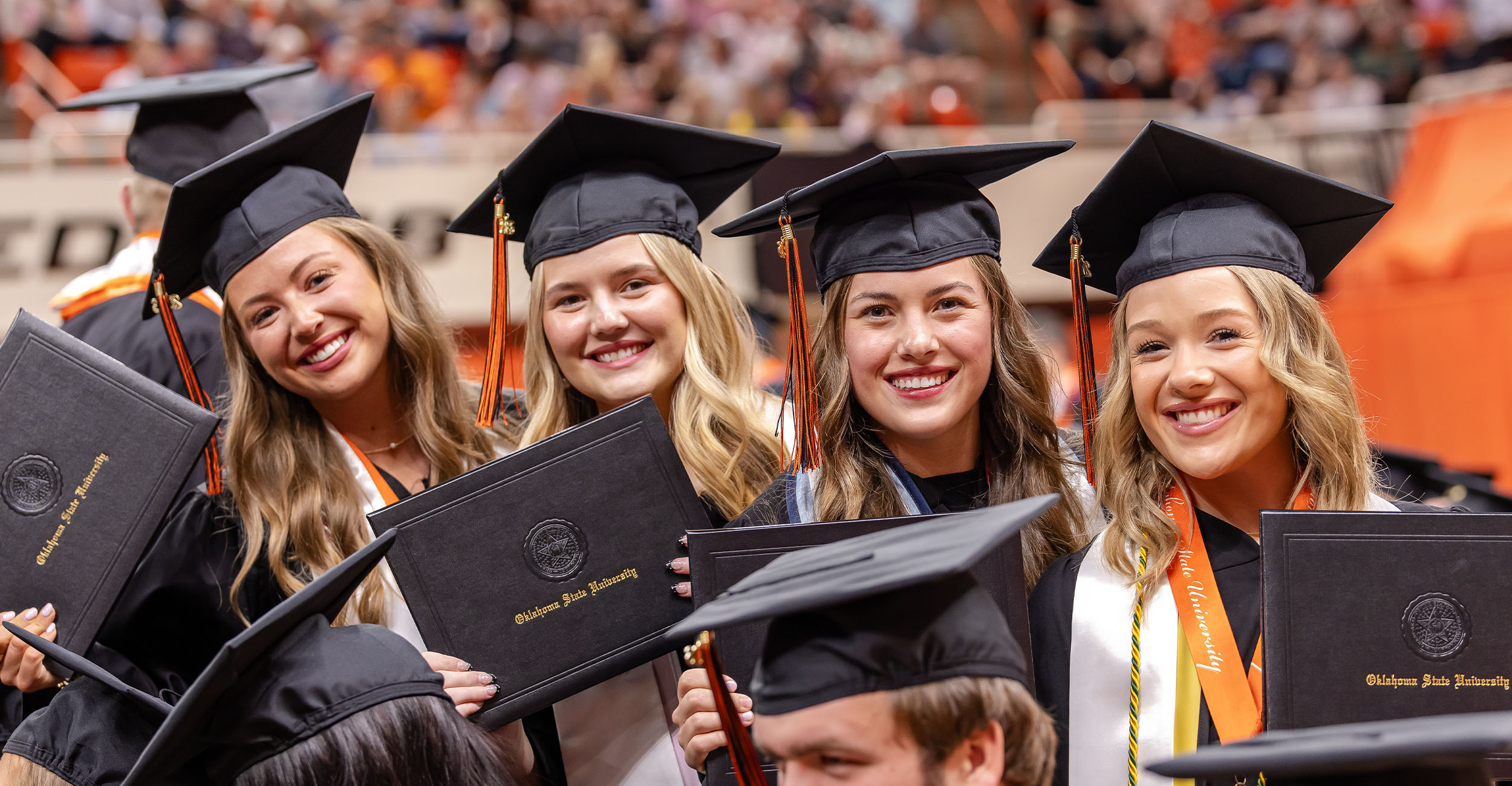 Four graduates in black caps and gowns hold Oklahoma State University diploma covers and pose together in a crowded arena during a commencement ceremony.