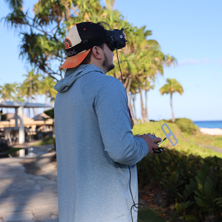 A person wearing FPV goggles and a backward black-and-orange cap holds a radio controller with antenna attachments while piloting a drone outdoors near palm trees and a coastal resort walkway.