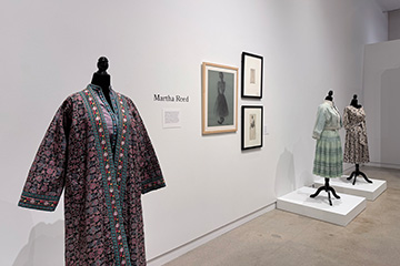 Exhibition gallery featuring three vintage garments displayed on mannequins alongside framed fashion sketches and interpretive text about designer Martha Reed.