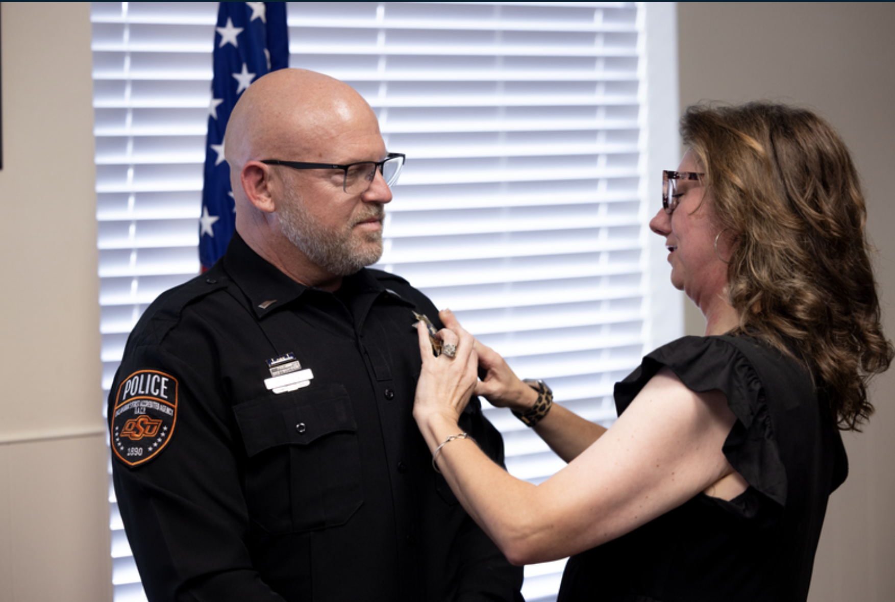 An OSU Police officer in uniform stands indoors while another person pins a badge or insignia onto the officer’s shirt during a ceremonial recognition event.