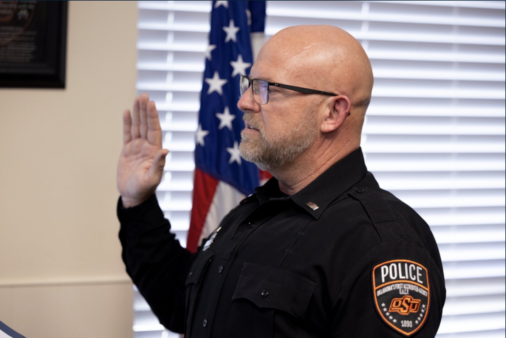 An OSU Police officer in uniform raises a right hand while taking an oath indoors, with an American flag visible in the background.