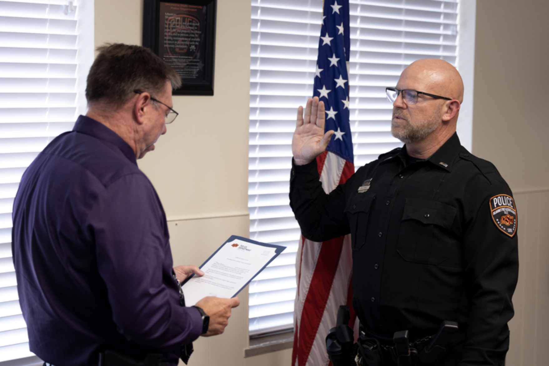 An OSU Police officer in uniform raises a hand while reciting an oath as another person reads from a document beside an American flag during a swearing-in ceremony.