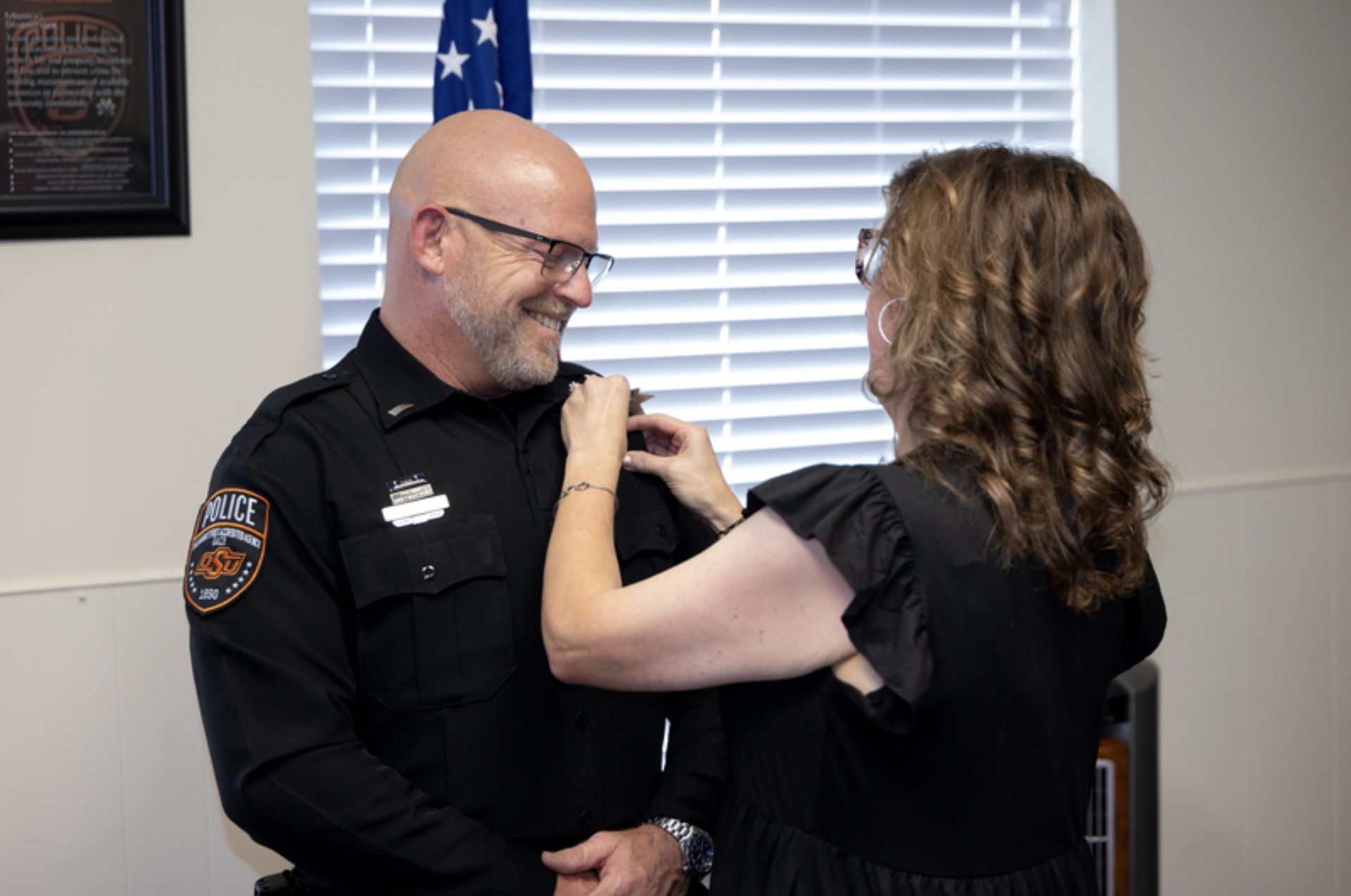 An OSU Police officer in uniform stands indoors while another person attaches an insignia to the officer’s collar during a promotion or recognition ceremony.