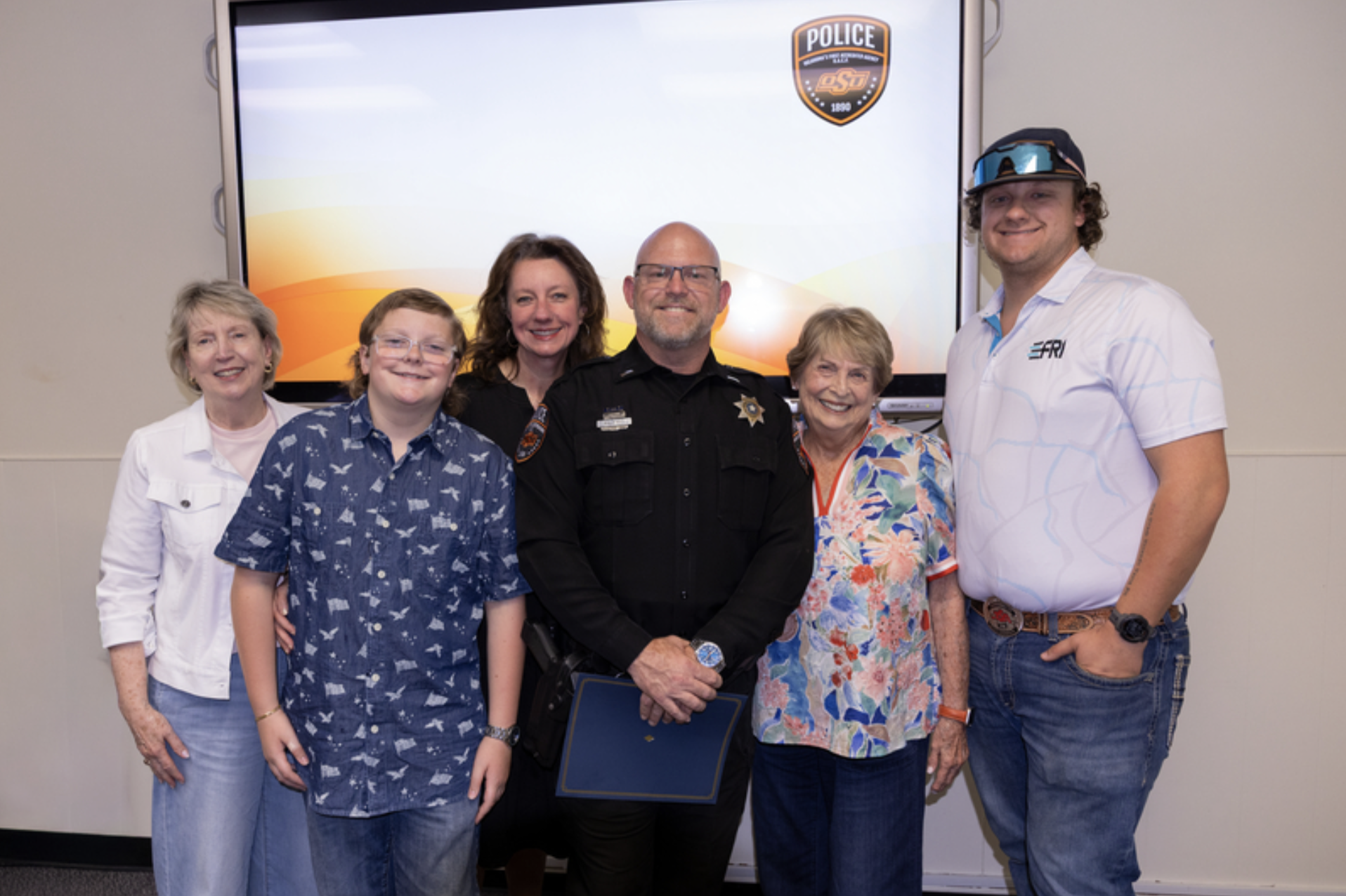 A uniformed OSU Police officer stands with five family members or supporters for a group photo during a recognition or promotion ceremony, with an OSU Police logo displayed on a screen behind them.