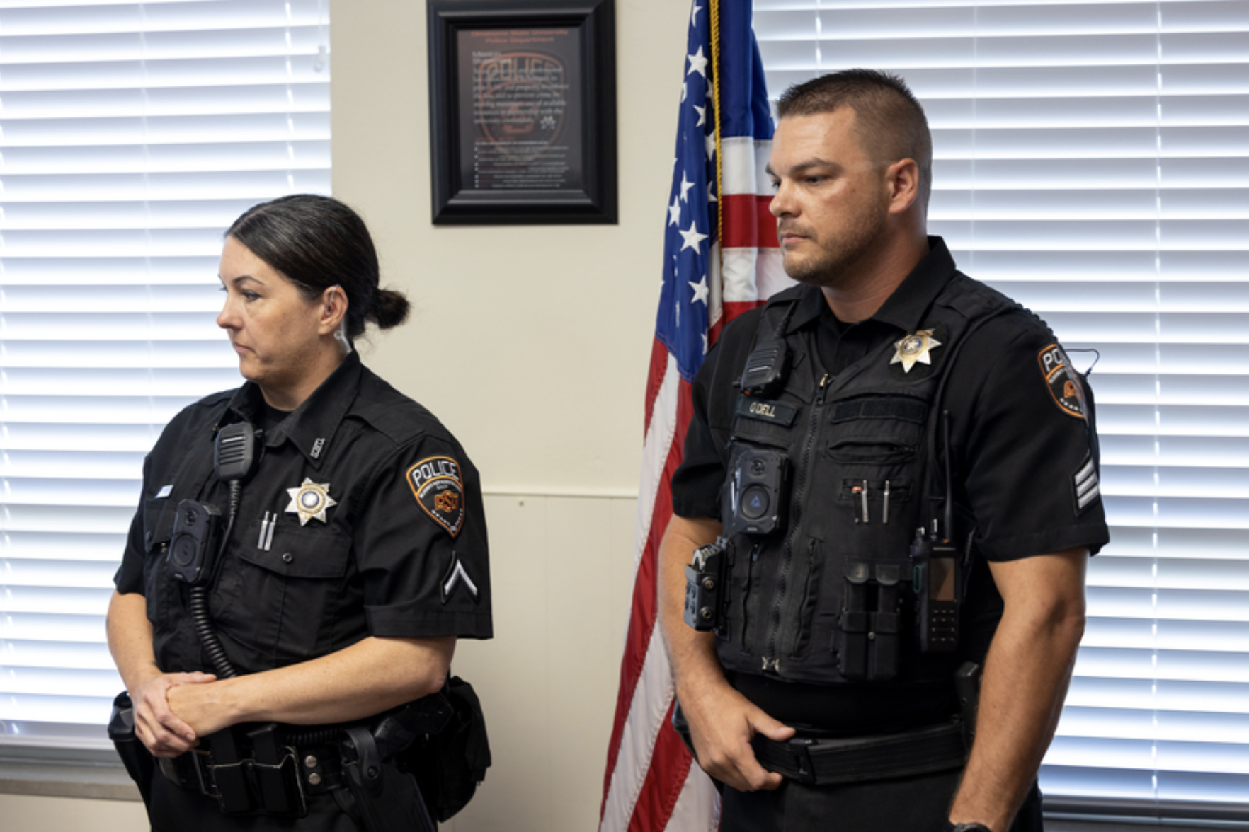 Two uniformed OSU Police officers stand in a meeting room near an American flag during a departmental recognition or ceremonial event.