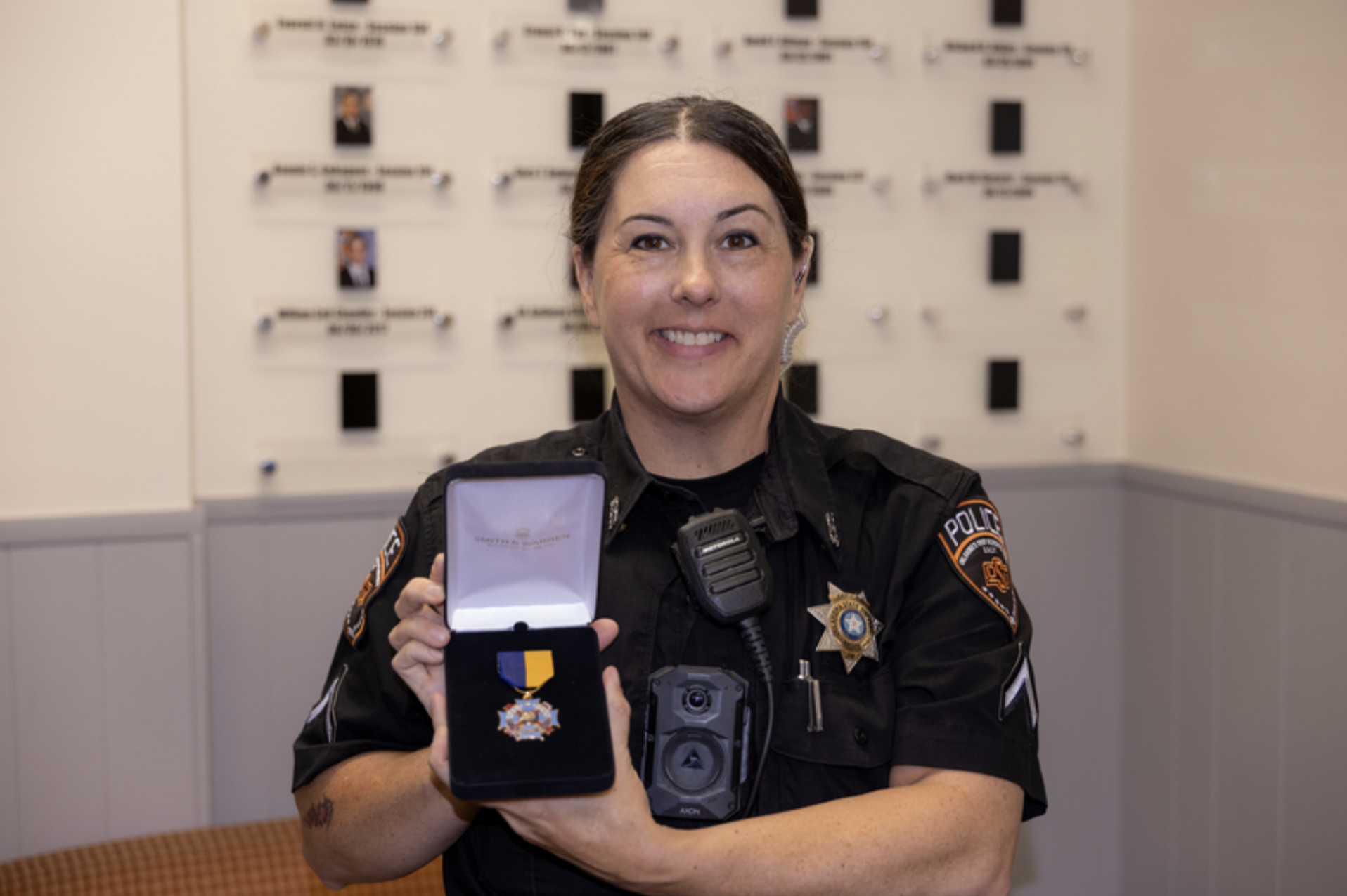 A uniformed OSU Police officer holds an open medal case displaying an award while posing for a portrait inside a police department office.
