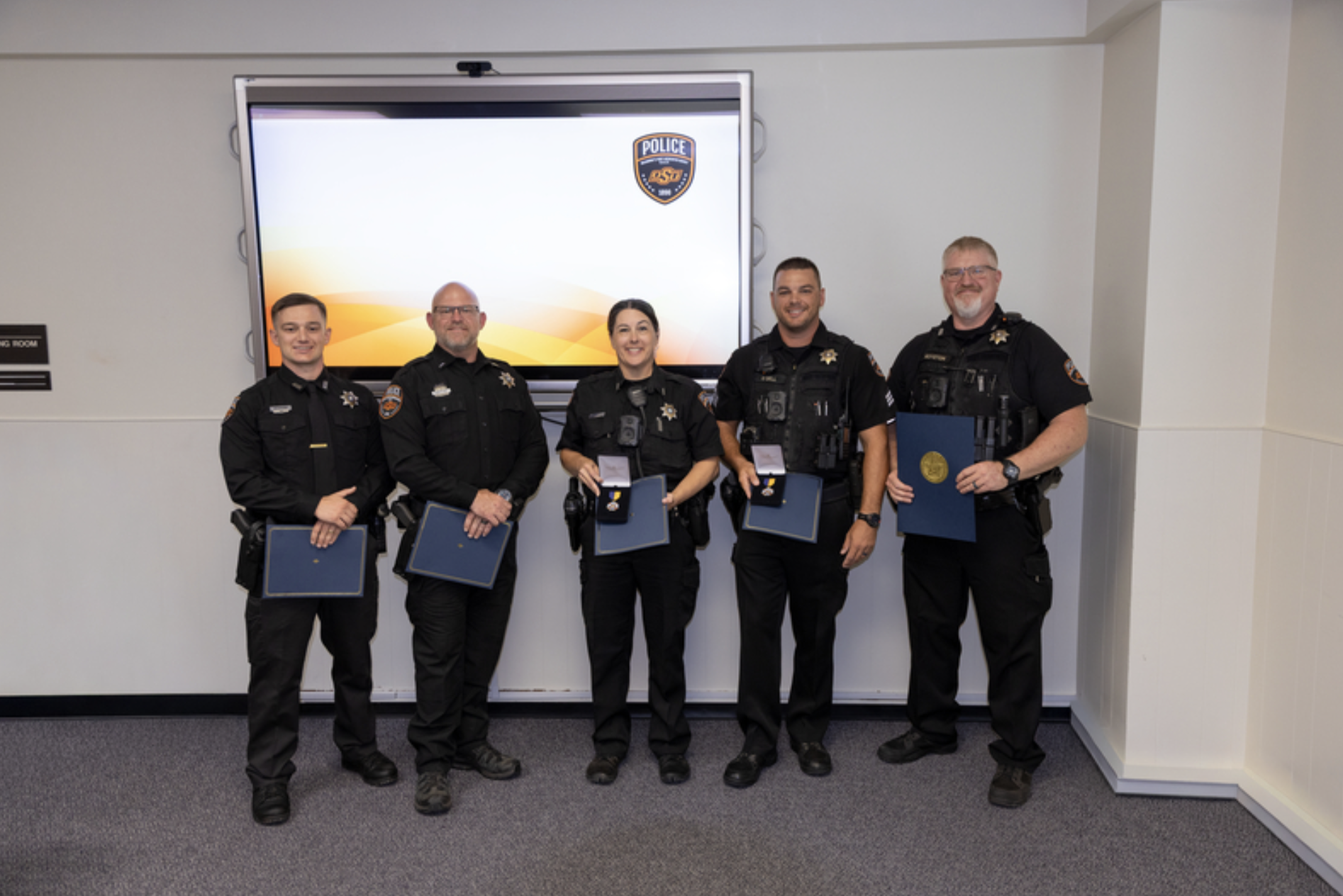 Five uniformed OSU Police officers stand together holding certificates and awards during a recognition ceremony, with an OSU Police logo displayed on a screen behind them.