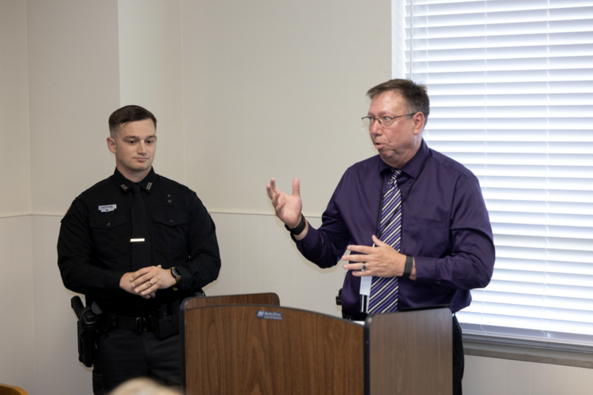 A uniformed OSU Police officer stands beside a person speaking at a podium during a recognition or awards ceremony in a meeting room.