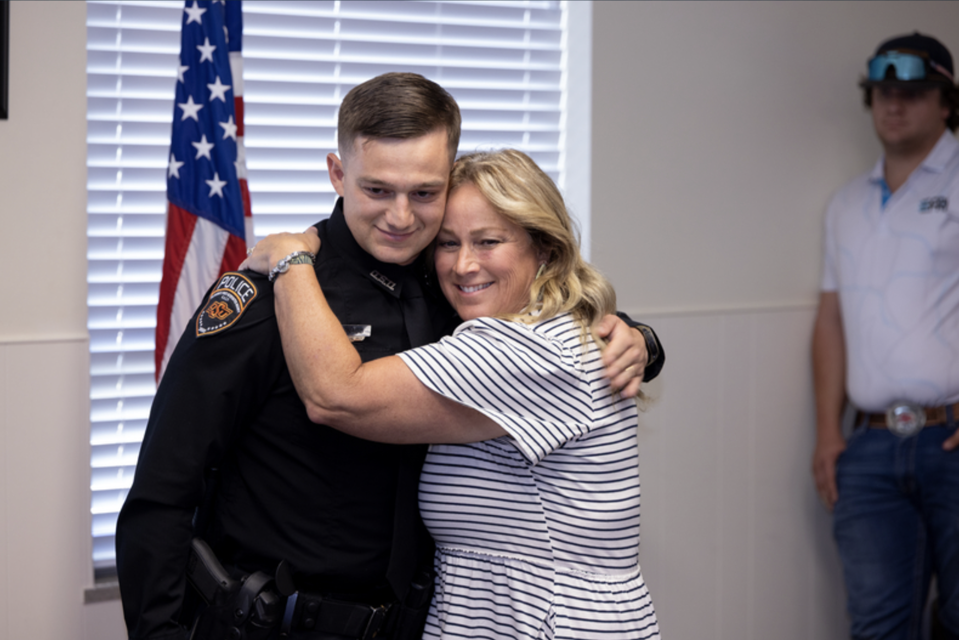 A uniformed OSU Police officer embraces an attendee during a recognition ceremony, with an American flag visible in the background.
