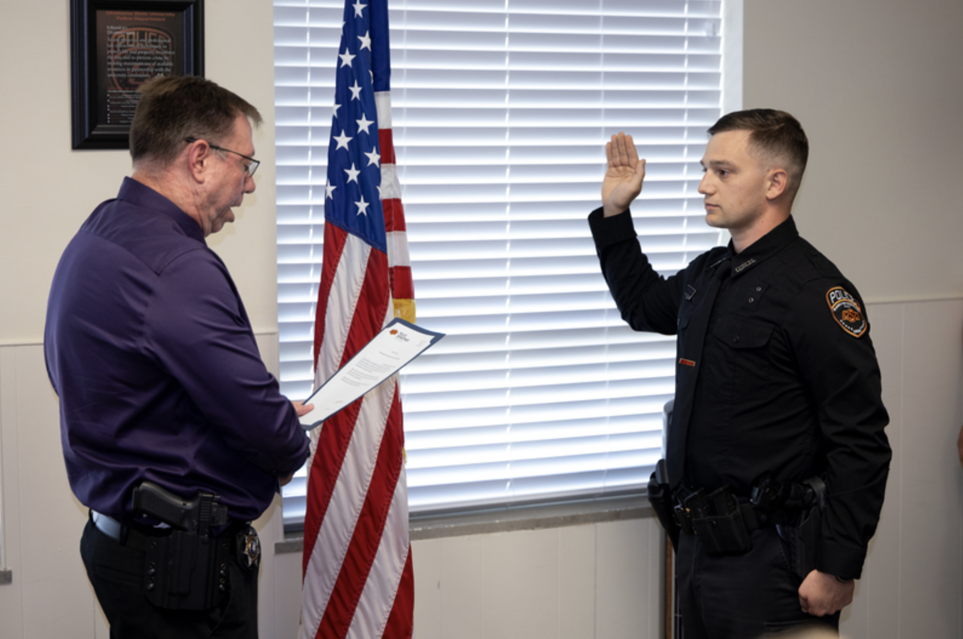 A uniformed OSU Police officer raises a hand while taking an oath as another person reads from a document beside an American flag during a swearing-in ceremony.