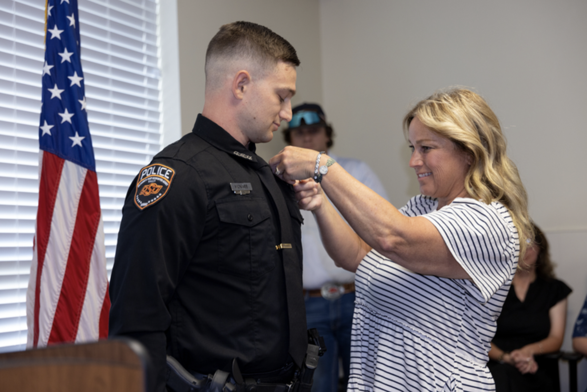 An attendee pins a badge or insignia onto the uniform of an OSU Police officer during a ceremonial recognition or swearing-in event, with an American flag visible nearby.