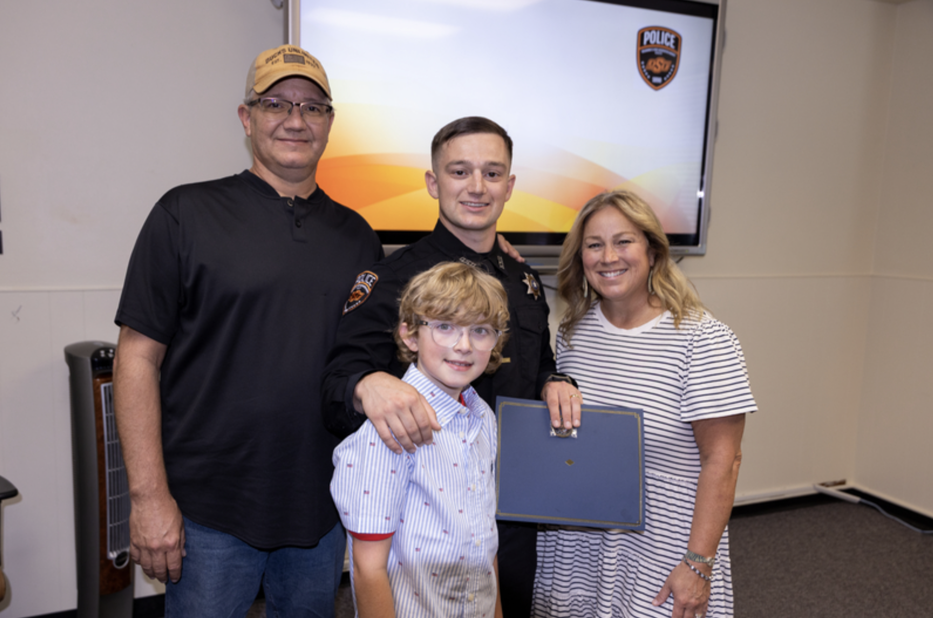 A uniformed OSU Police officer stands with three family members while holding a certificate during a recognition ceremony, with an OSU Police logo displayed on a screen behind them.