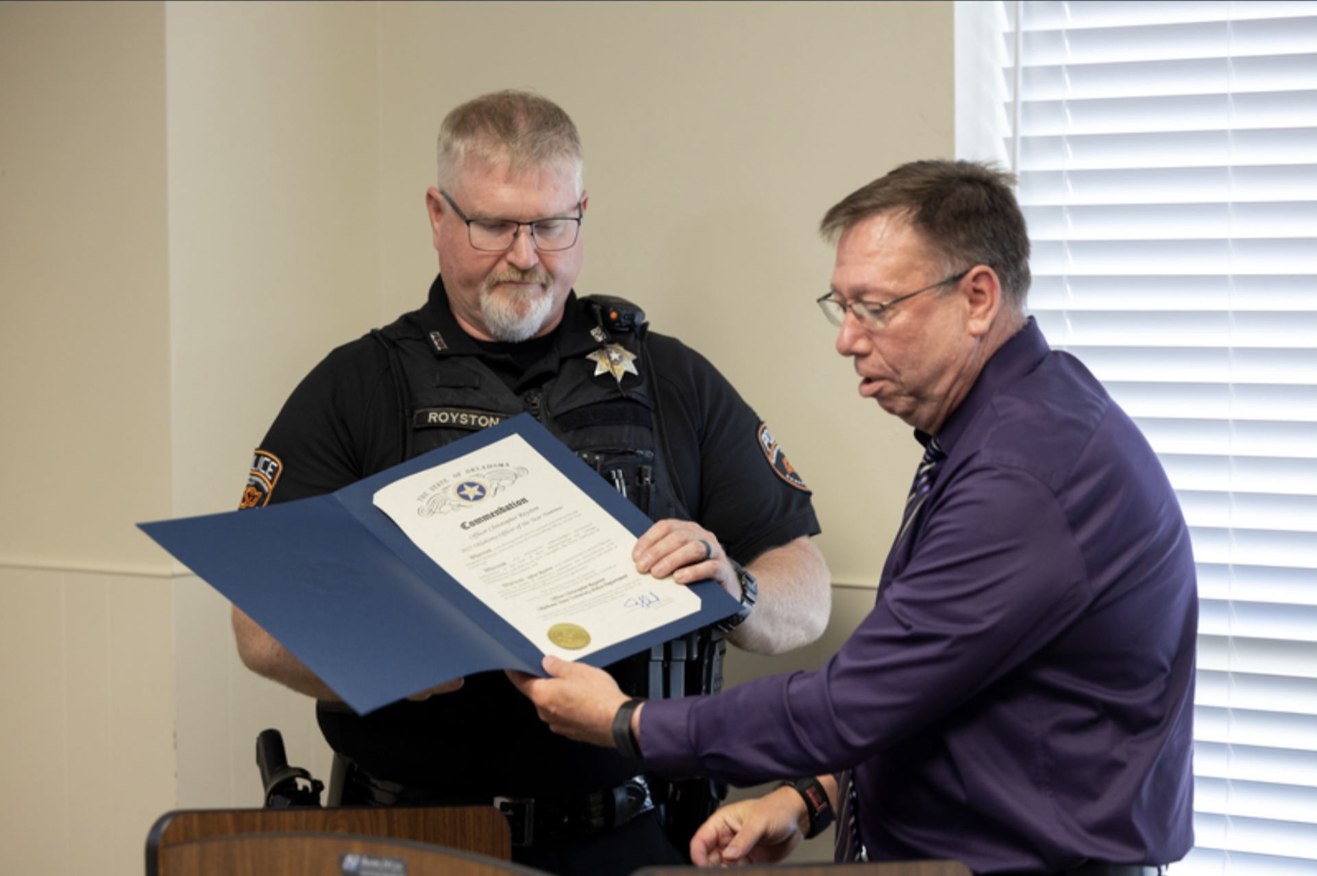 A uniformed OSU Police officer receives a certificate of commendation from a presenter during a recognition ceremony in a meeting room.