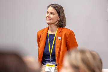 A person wearing an orange blazer and a name badge labeled “DIRECTOR Rachael Eaton” stands at the front of a room near a speaker, addressing a seated audience during a formal meeting or presentation.