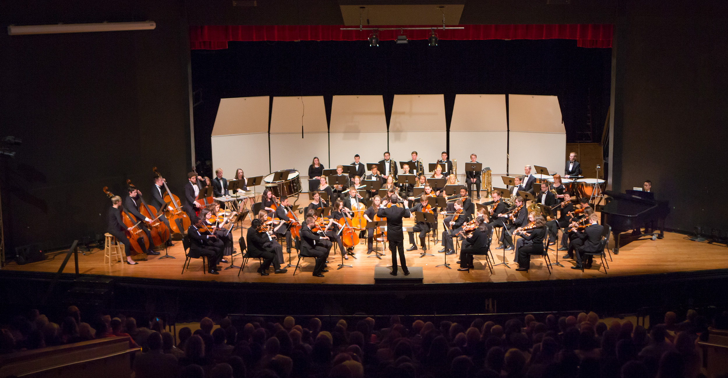 A large orchestra performs on a concert hall stage, with a conductor standing at center facing seated musicians and an audience visible in the foreground.